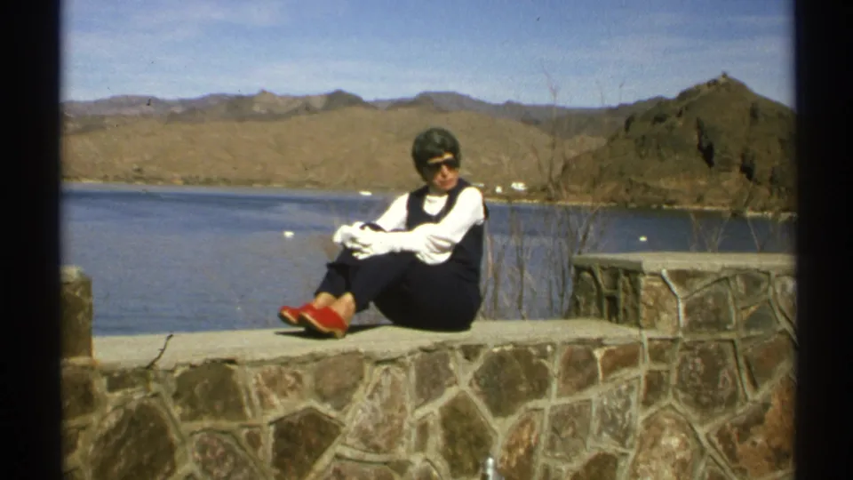 The woman remains seated on the stone wall in Arizona, with lake water, dry hills, and rocky desert terrain behind her.