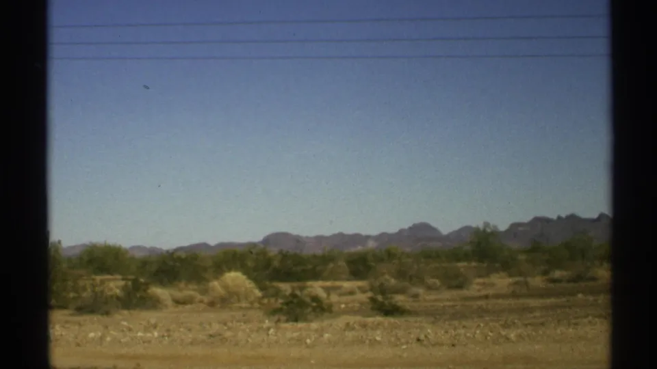 1972 Arizona desert landscape with dry ground, shrubs, faint power lines, blue sky, and mountains viewed from a moving vehicle.