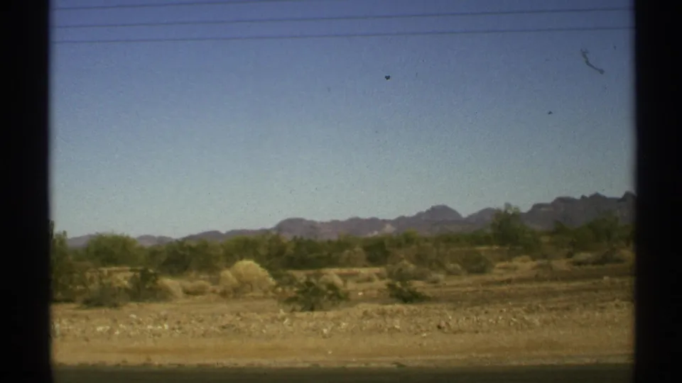 Arizona desert scenery in 1972 with sparse shrubs, sandy ground, overhead power lines, and rugged mountains beyond the road.