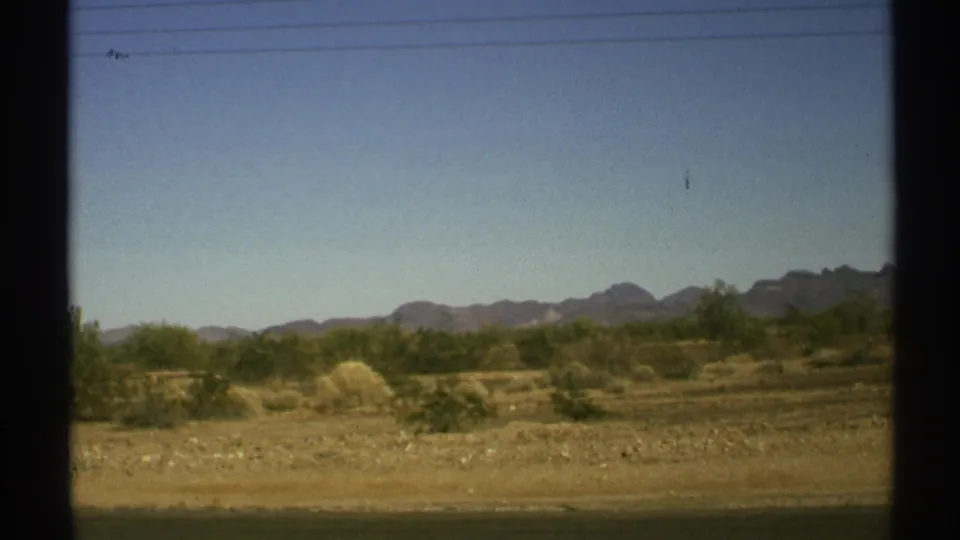 A moving-vehicle view of 1972 Arizona desert ground, scattered shrubs, faint utility lines, and low mountains under clear sky.