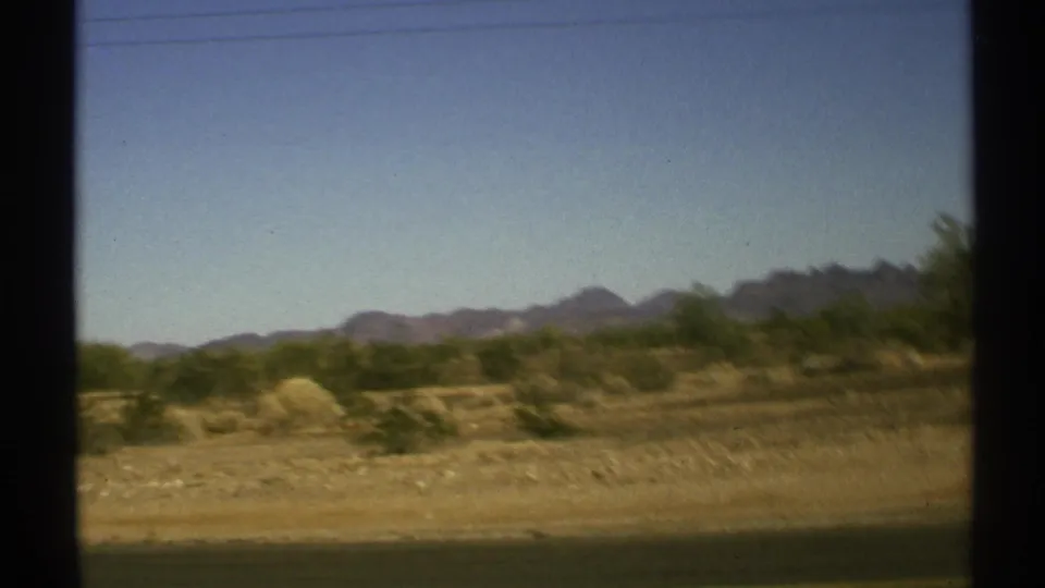 1972 Arizona desert roadside view with scrub, distant mountains, pale sky, and dark window framing from a moving vehicle.