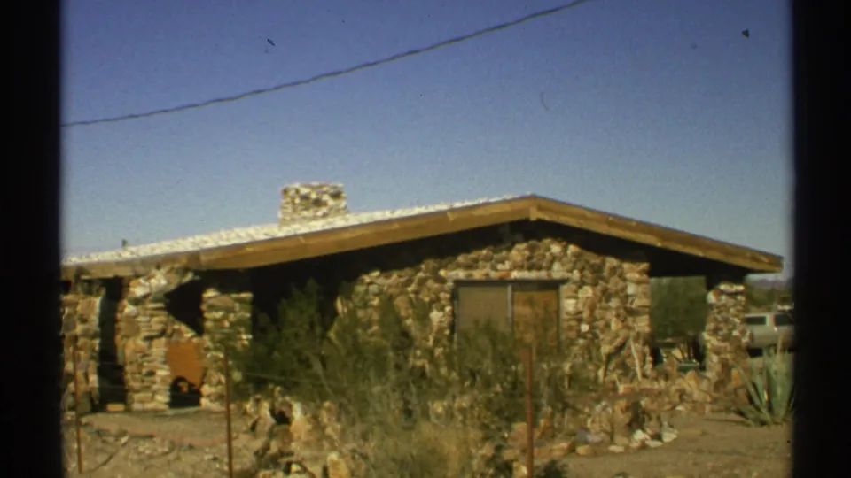1972 Arizona 8mm frame showing a low stone house with a chimney, desert shrubs, yard fencing, and clear blue sky.