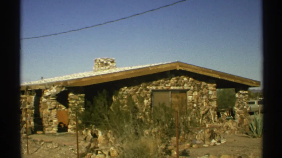 1972 Arizona 8mm frame showing the stone house, chimney, desert vegetation, fence posts, and a partial vehicle at the right edge.