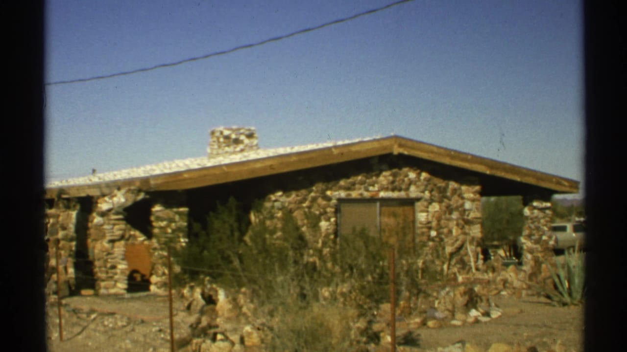 Stone desert building behind a wire fence with scrub and blue sky.