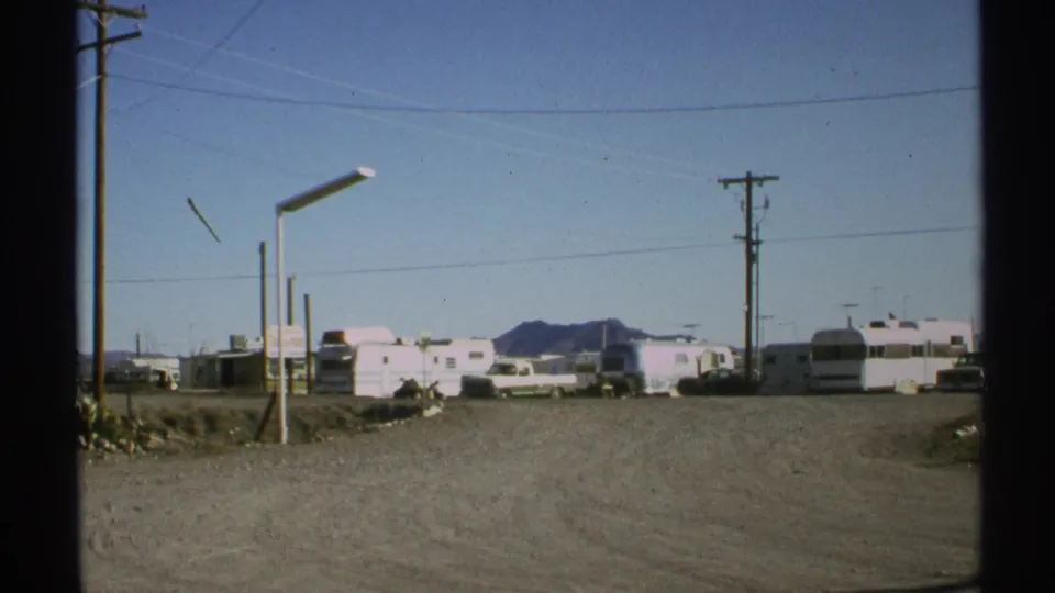 1972 Arizona campground view with RVs and trailers on dusty ground, utility lines crossing the sky, and mountains in the background.