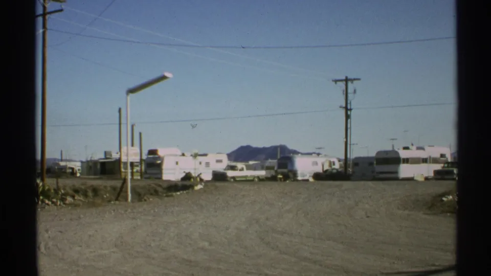 1972 Arizona 8mm still of a broad dusty campground road, parked trailers, an RV, utility poles, and distant mountains.