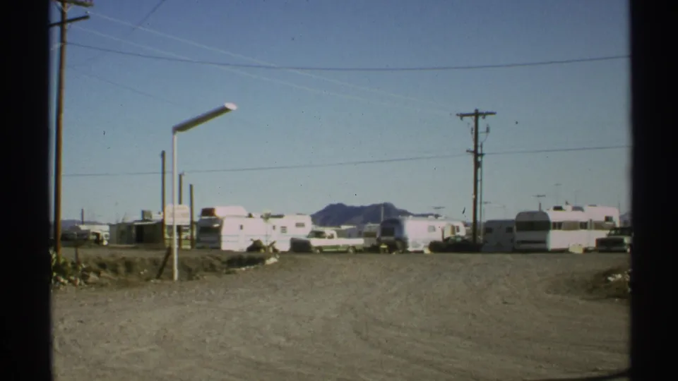 1972 Arizona desert campground frame with white trailers, RVs, utility poles, overhead wires, dusty ground, and low mountains.