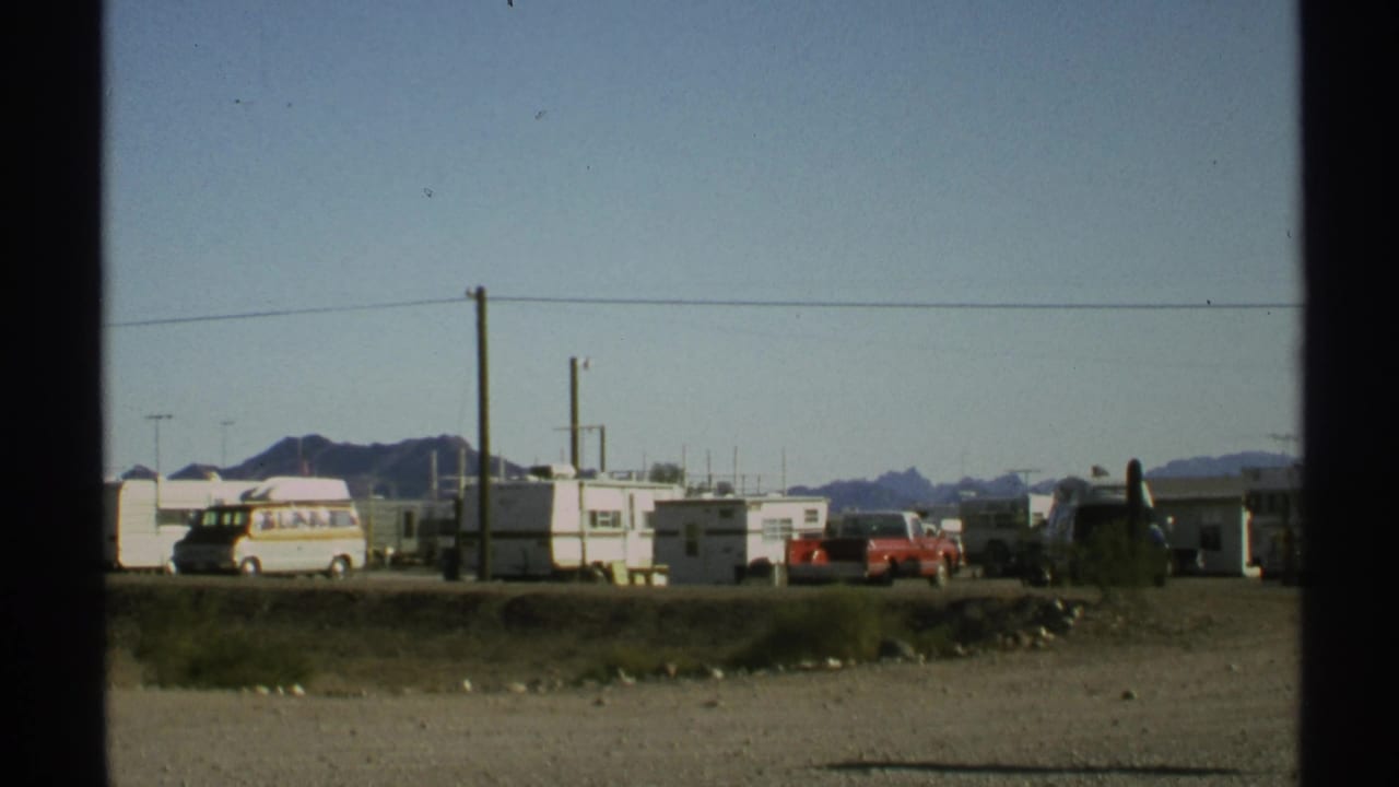 Desert RV park with trailers, vehicles, utility poles, power lines, and distant mountains.