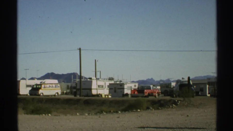 1972 Arizona 8mm frame showing trailers and parked vehicles beside a gravel desert roadside with utility poles and mountains beyond.