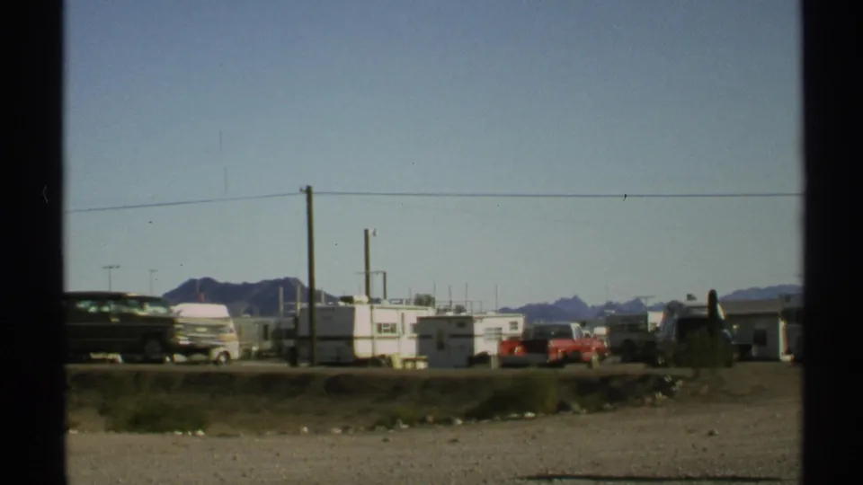 1972 Arizona roadside view with a dark station wagon moving past white trailers, a red pickup, utility poles, and distant desert mountains.