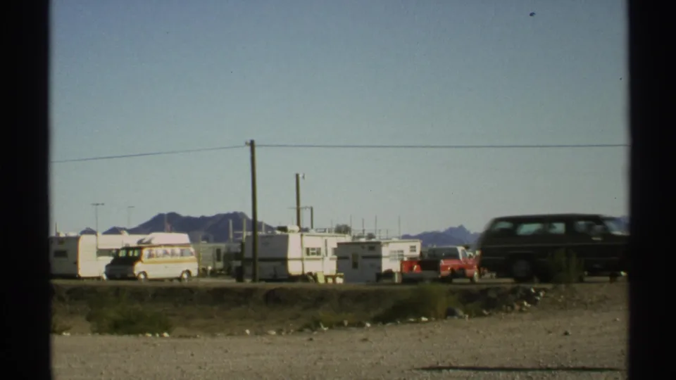 1972 Arizona 8mm frame showing a dark station wagon passing an RV park with trailers, a red pickup, power lines, and low mountains.