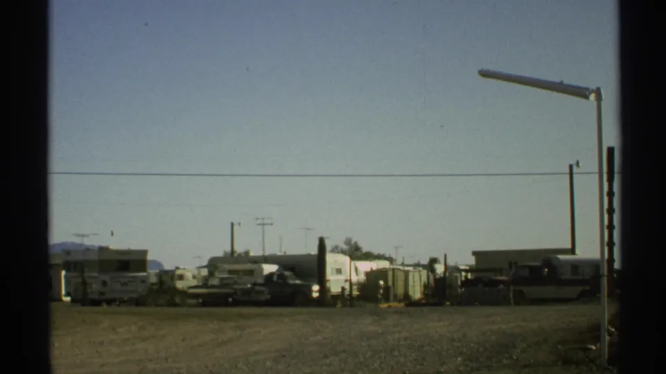 1972 Arizona desert roadside frame showing a gravel approach to an RV park with trailers, parked cars, utility poles, and a low structure.