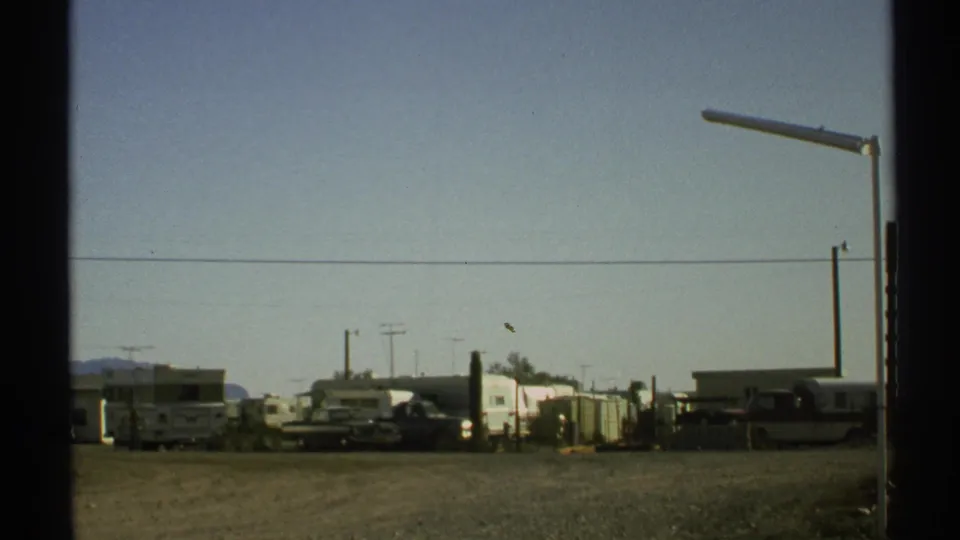1972 Arizona roadside view with parked RVs, trailers, low buildings, utility poles, and distant mountains.