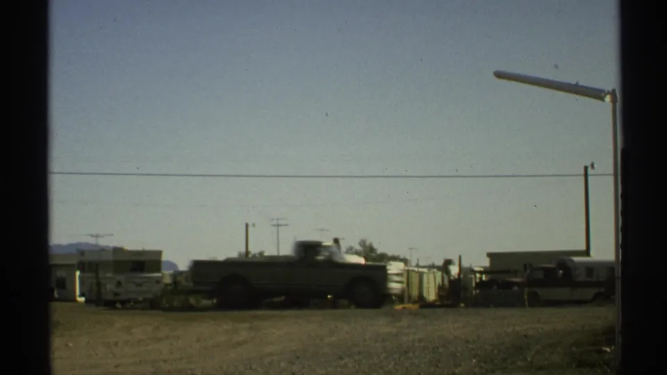 A pickup truck crosses the foreground of a 1972 Arizona roadside scene with RVs, trailers, and utility poles behind it.