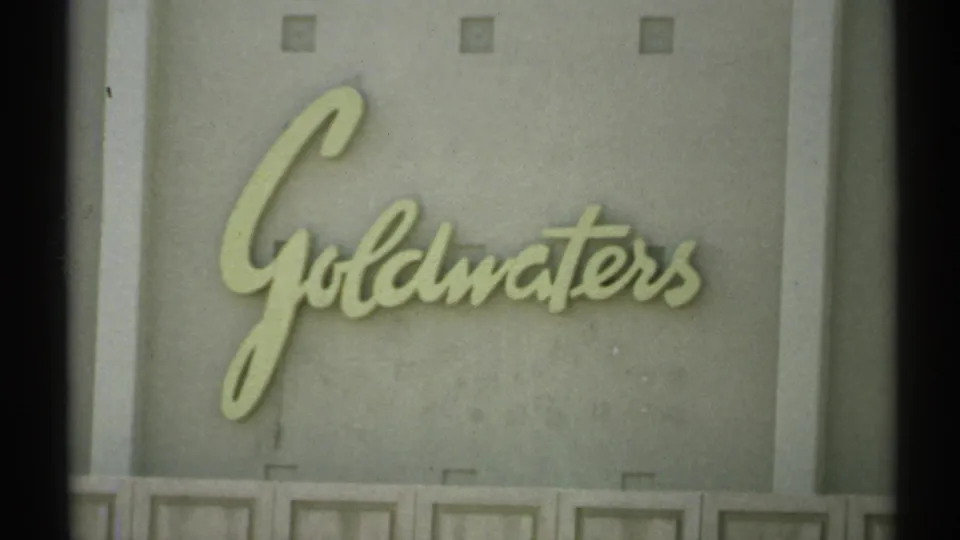 Handheld 8mm view of a pale gray wall with raised cursive Goldwaters lettering, likely part of an indoor display or storefront setting.
