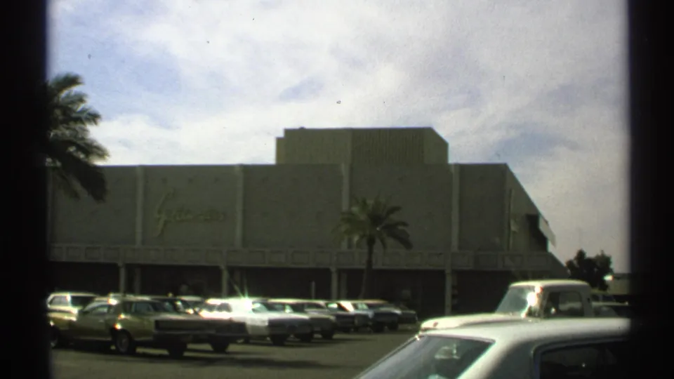 Wide 1972 Arizona home-movie frame of Goldwaters across a sunlit parking lot with period cars and palms.
