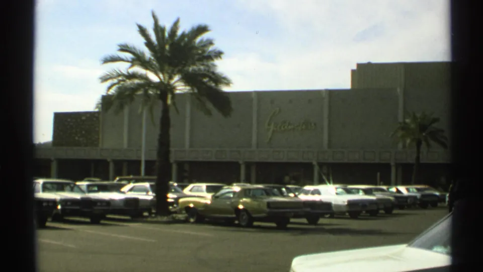 1972 Arizona 8mm frame with palm landscaping, rows of parked cars, and a large signed commercial building across the lot.