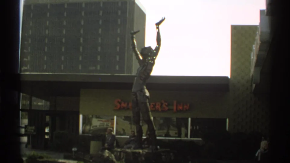 1972 Arizona home-movie frame showing a raised-arm statue in front of Smokey's Inn, with seated visitors and a tall modern building behind.