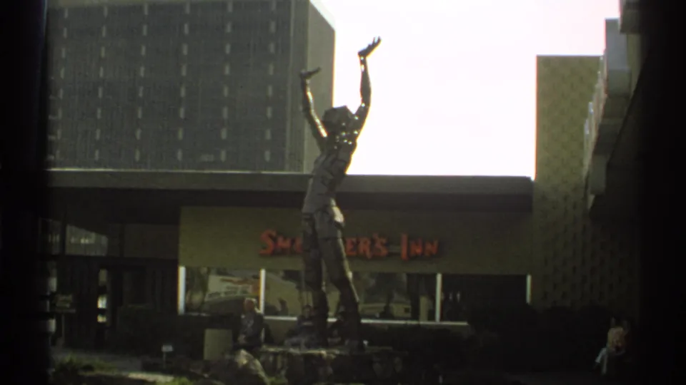 The statue remains centered outside Smokey's Inn, with people seated near the entrance and a tall building filling the background.