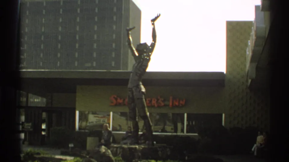 A closer 8mm view frames the raised-arm statue against the Smokey's Inn facade, with a tall building and seated people still visible.