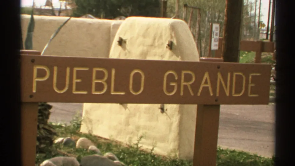 1972 Arizona frame showing a wooden Pueblo Grande sign near a road, with a low light-colored wall and surrounding landscaping.