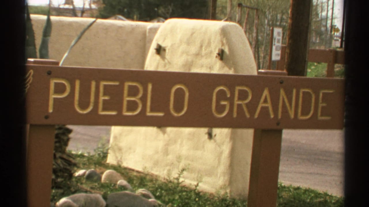 Wooden sign reading PUEBLO GRANDE beside a stucco wall and roadside landscaping.