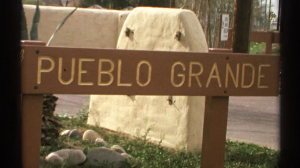 Vintage 8mm frame of the Pueblo Grande sign in Arizona, with gold lettering, desert landscaping, and adobe-style site walls.