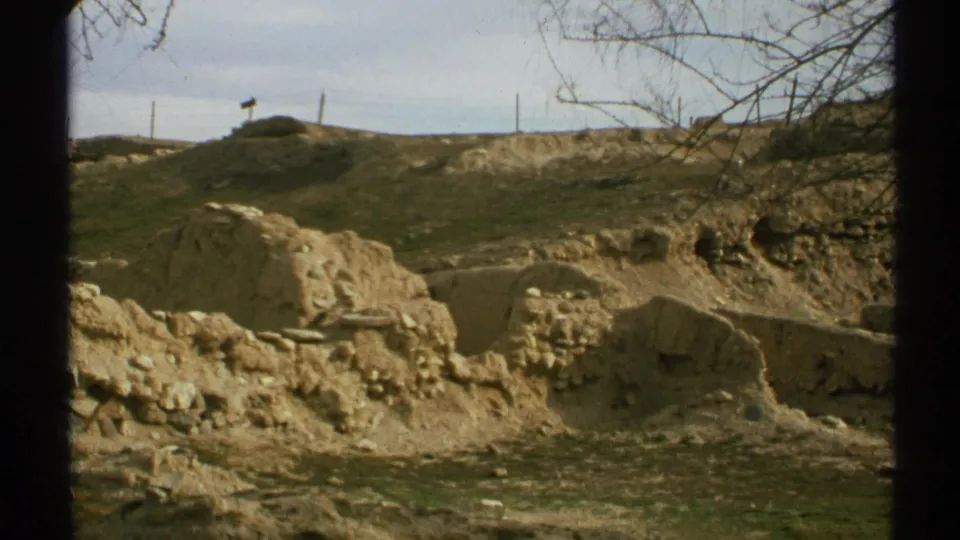 1972 Arizona 8mm view of low adobe-like remains, exposed dirt, rocks, a grassy mound, and utility poles in the distance.