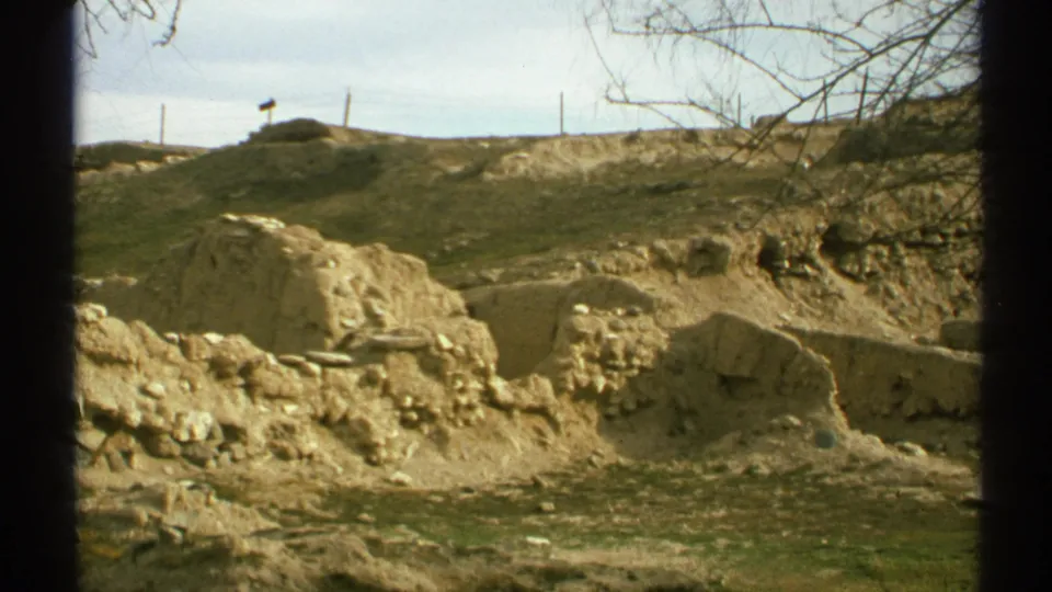 1972 Arizona home-movie frame showing adobe-like ruin remains, rough dirt, sparse grass, bare branches, and a low desert hillside.