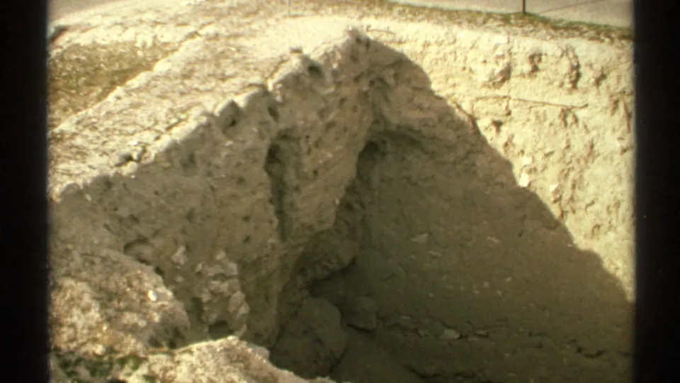 1972 8mm Arizona frame showing a tan trench or excavation pit, rough soil walls, shadowed earth, and sparse desert ground nearby.