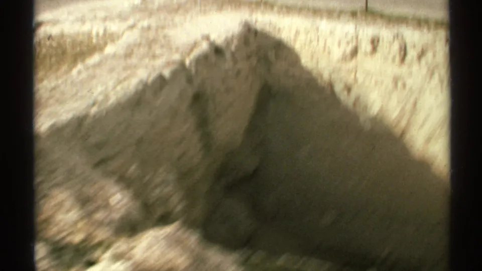 Soft handheld 8mm view of a tan excavation wall with bright sunlight and deep shadow across the trench.