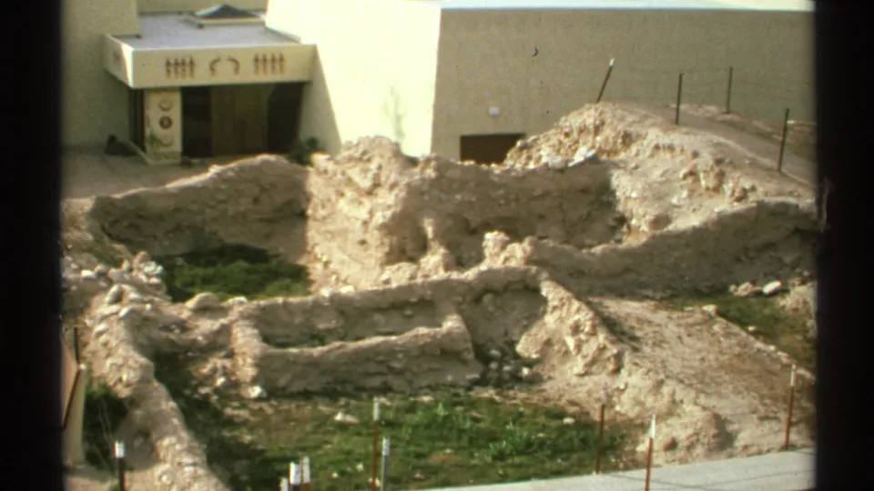 1972 Arizona frame showing low tan foundation walls, green ground patches, stakes, and a white building with decorative entrance details.