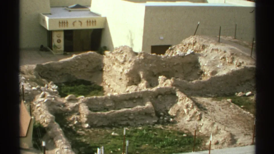 Handheld 8mm view of tan archaeological foundations and trench lines in front of a white building in Arizona, 1972.