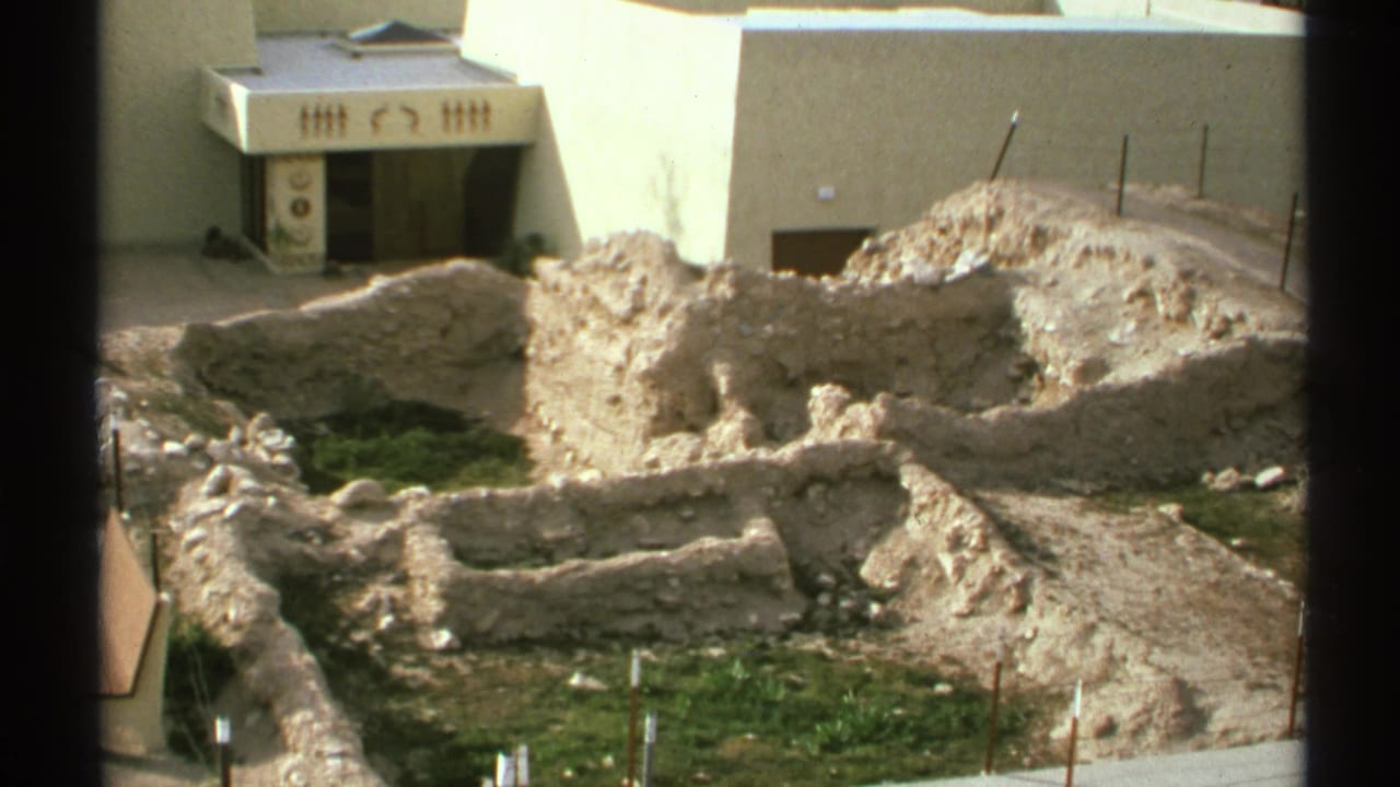 1972 Arizona 8mm frame showing pale excavation trenches, low ruins, grass patches, and a white building with Egyptian-style markings.