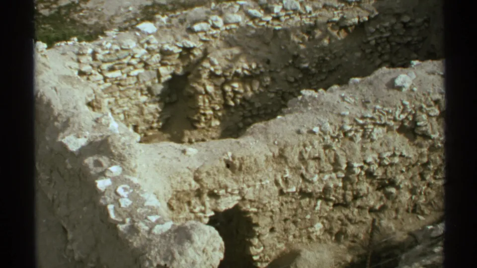 1972 Arizona 8mm frame with weathered ruin walls, a pale trench, scattered stones, and dark vertical bars crossing the foreground.