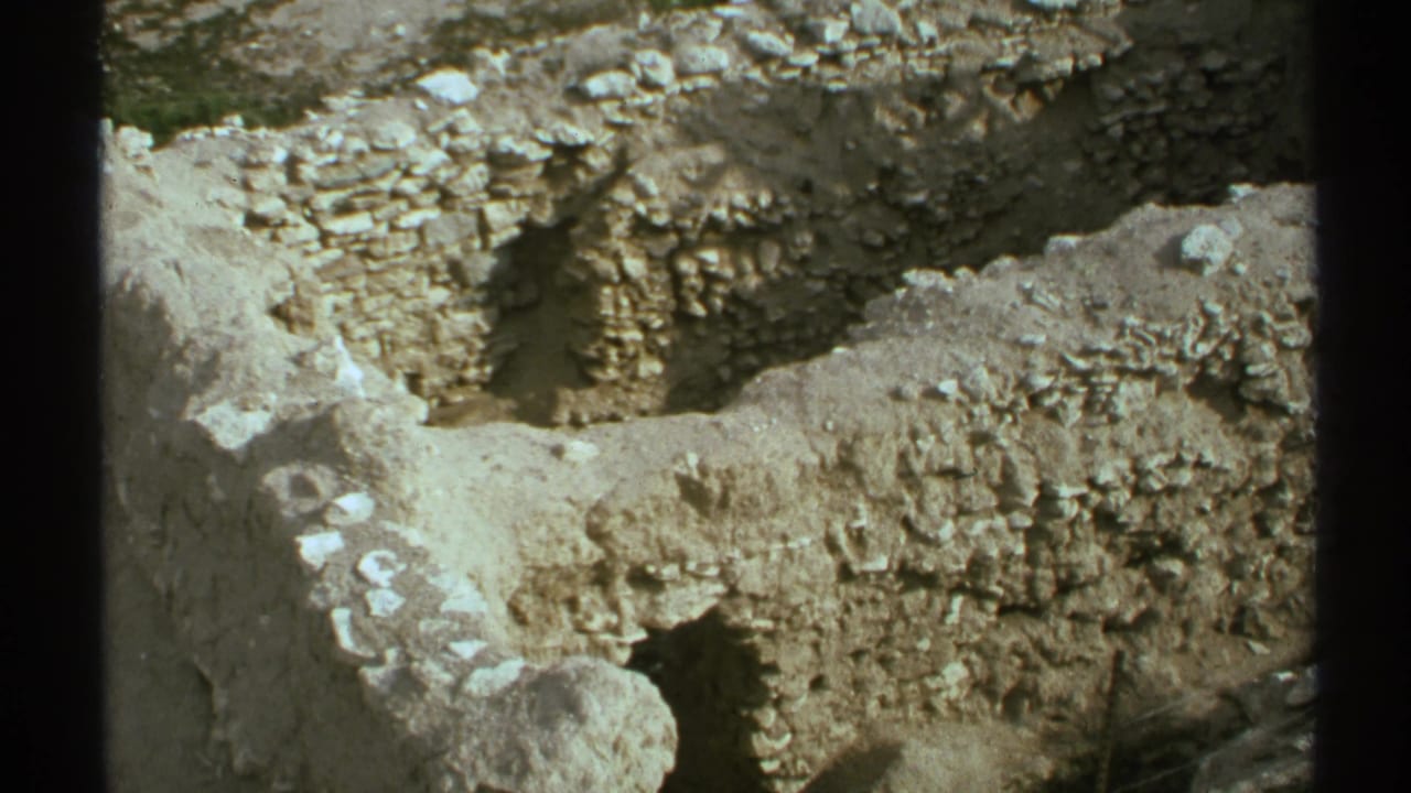 Sunlit stone ruin walls and rubble seen through dark vertical foreground bars.