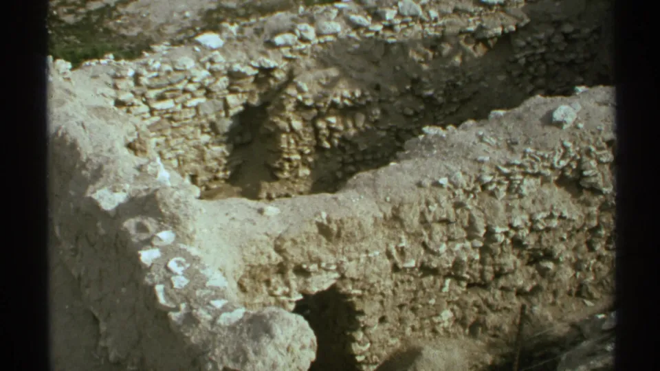 1972 Arizona home-movie frame looking down at exposed stone ruin walls, pale soil, and scattered rubble through dark vertical foreground bars.
