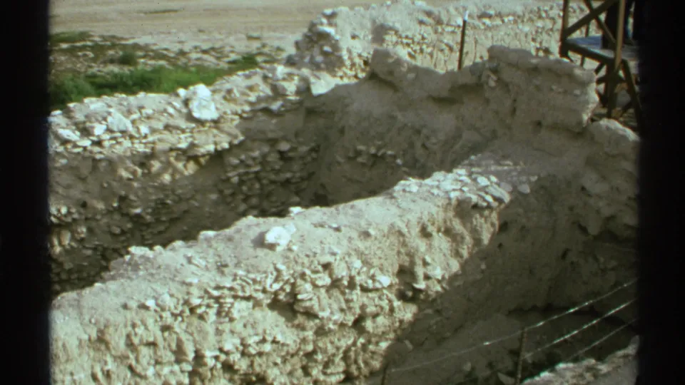 1972 Arizona home-movie frame of exposed stone ruin walls, loose rubble, dark foreground framing, and metal fencing along the lower right edge.
