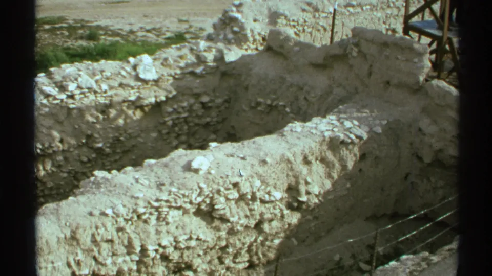 1972 Arizona excavation frame looking over pale stone foundations, debris, a dark foreground bar, and protective fencing near the lower right.