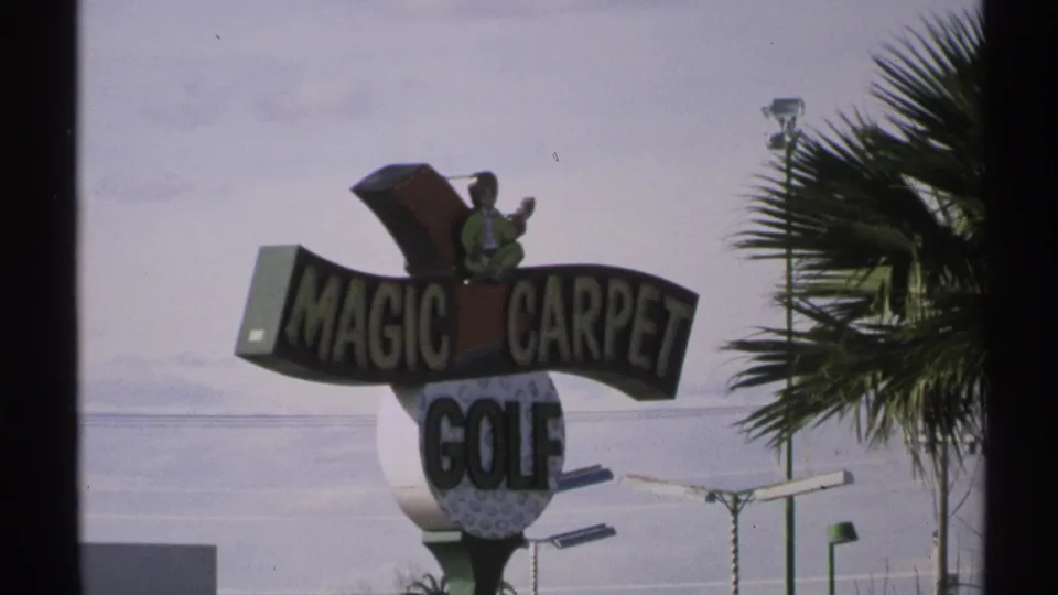 Magic Carpet Golf sign in Tucson-area 1972 home-movie footage, with a golfer figure, palm fronds, street lights, and pale sky.