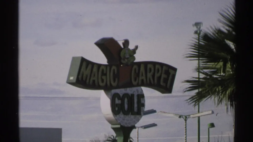 Second view of the Magic Carpet Golf sign, showing the attraction name, golfer figure, palm fronds, sky, and roadside lighting.