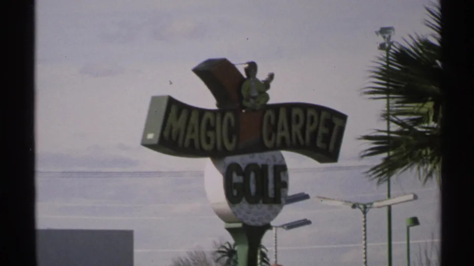 1972 Arizona 8mm frame showing a Magic Carpet Golf sign near a road, with palm foliage and a faded sky in the background.