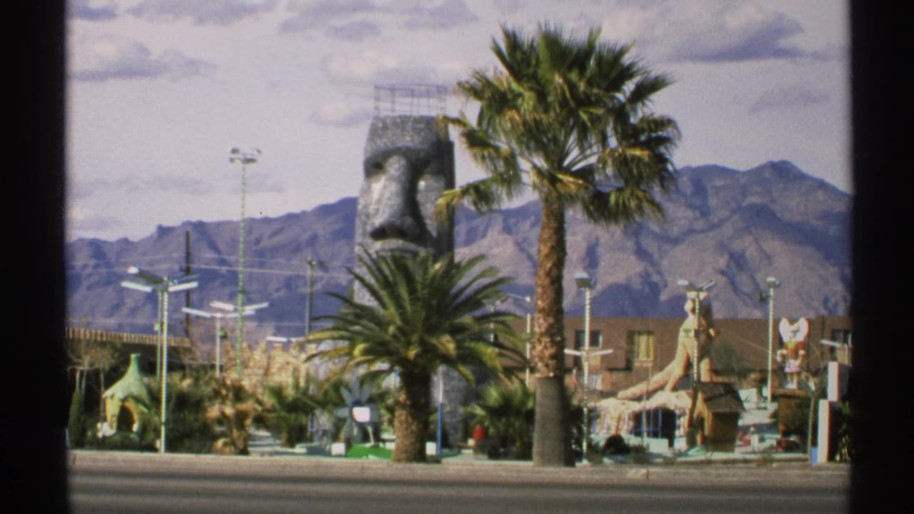 Large stone face sculpture, palm trees, themed structures, road, and mountains.