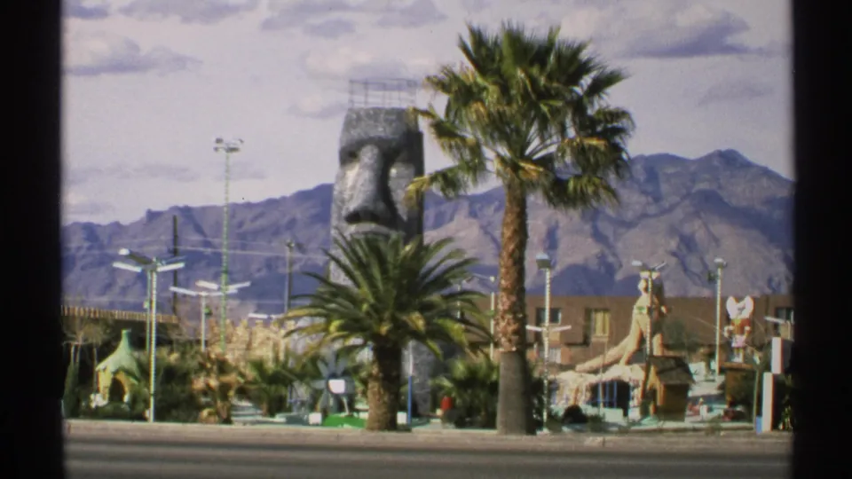 1972 Arizona roadside attraction with a giant stone face sculpture, palms, fantasy structures, and mountains beyond the road.