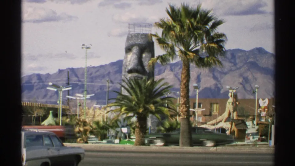 1972 Arizona 8mm view of the attraction from across the road, with palm trees, a stone face sculpture, mountains, and cars passing.