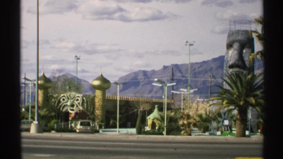 1972 Arizona frame showing whimsical roadside attraction buildings, a partial stone face sculpture, palm trees, road, and desert mountains.
