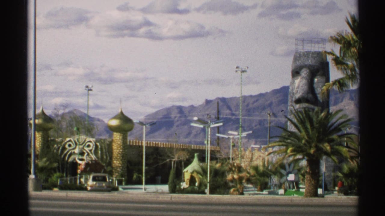 Roadside attraction with golden domes, tiger-head sculpture, palm tree, parked car, and desert mountains.