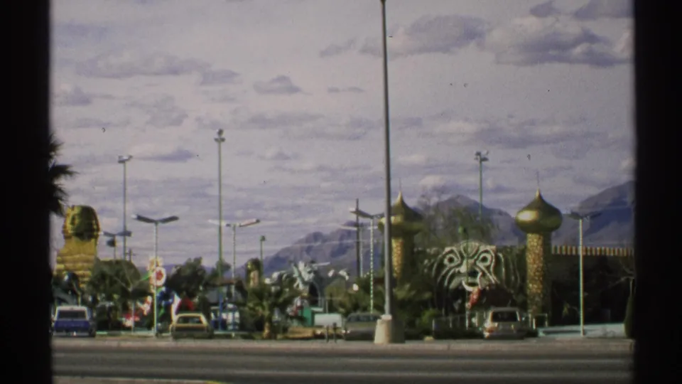 Wide 1972 Arizona 8mm frame of the roadside attraction, showing domes, a tiger-head sculpture, cars, light poles, and desert mountains.