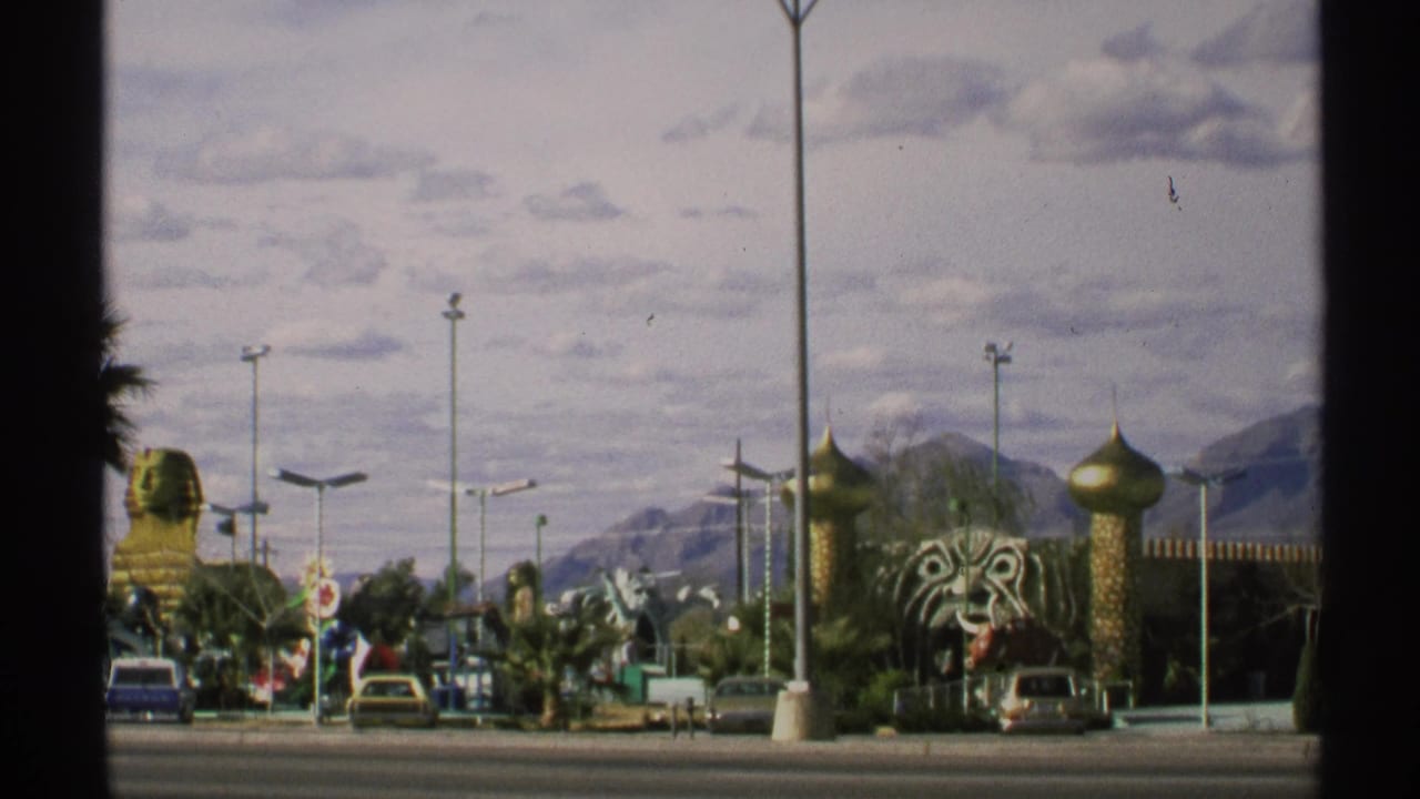 1972 Arizona home-movie frame showing a roadside attraction area with decorative facades, cars, lamp posts, desert landscaping, and mountains beyond.