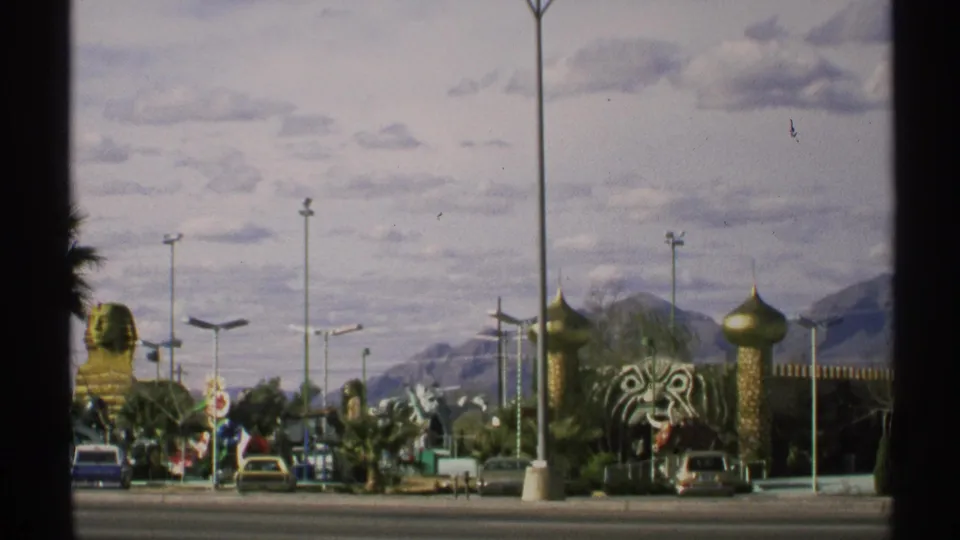 1972 Arizona home-movie frame showing a roadside attraction area with decorative facades, cars, lamp posts, desert landscaping, and mountains beyond.